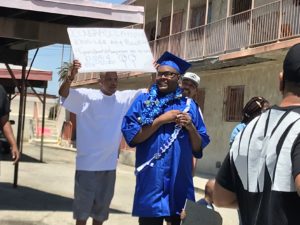  Man holding up handmade sign congratulating a smiling student in graduation cap and gown in front of him.