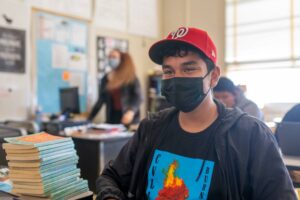 NMSS student in red baseball hat, mask, and black tshirt and jacket sits in classroom while teacher behind him gives instruction.