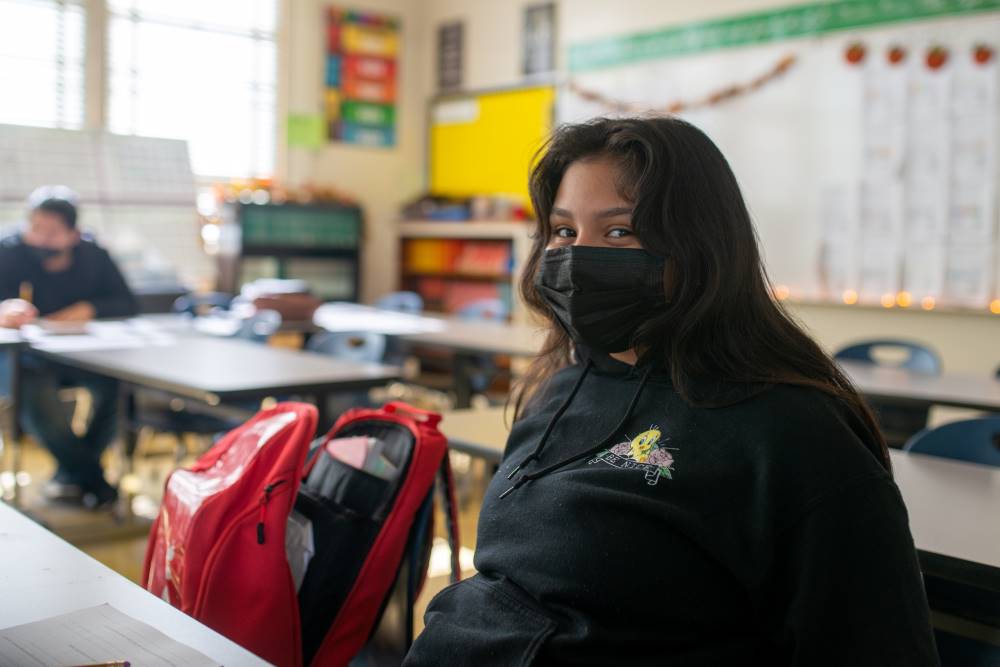 NMSS student sits at a school desk with a mask on and an open red backpack next to her.