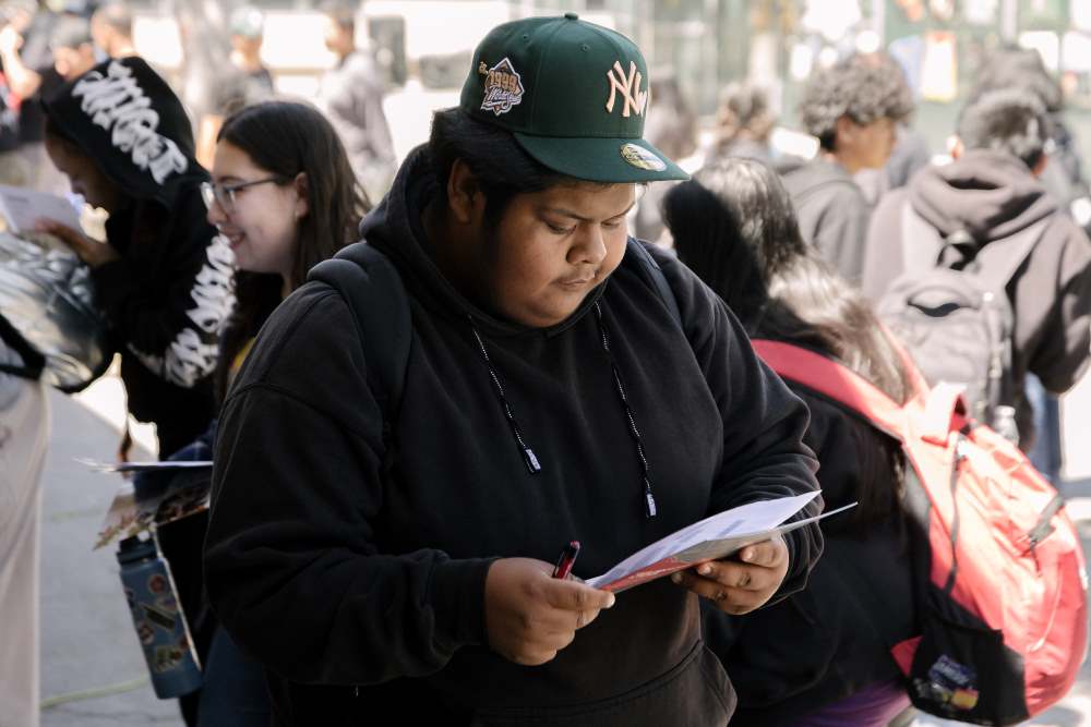 NMSS high school student in green hat looks intently at papers in his hand while other students mingle behind him outside.
