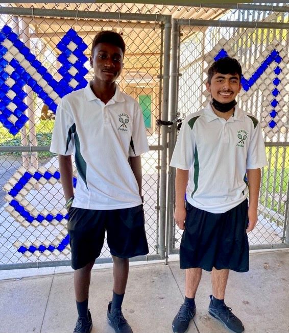 Two NMSS students in Gardena High white polo shirts and dark shorts tennis uniforms stand in front of NMSS gates.