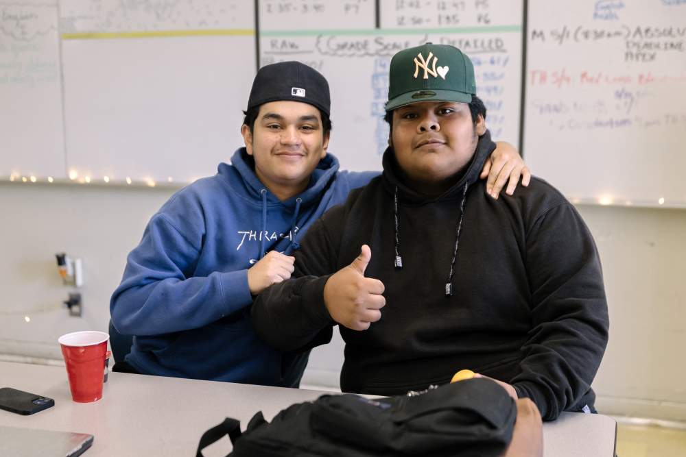 Two NMSS students with baseball caps on sit at a classroom table smiling and one giving a thumbs up.