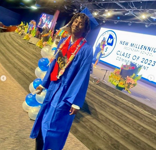 NMSS student in graduation cap and gown smiles as they walk down steps with a “Class of 2023” sign behind them on a stage.