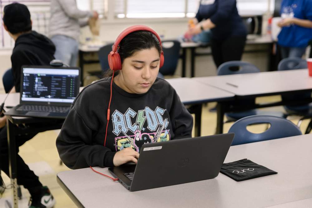 NMSS students sits at classroom table typing on laptop and wearing red headphones.