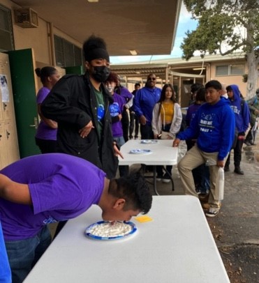 NMSS students bend over table with plate of whip cream and attempt to eat it with their hands tied behind their backs while other students look on.