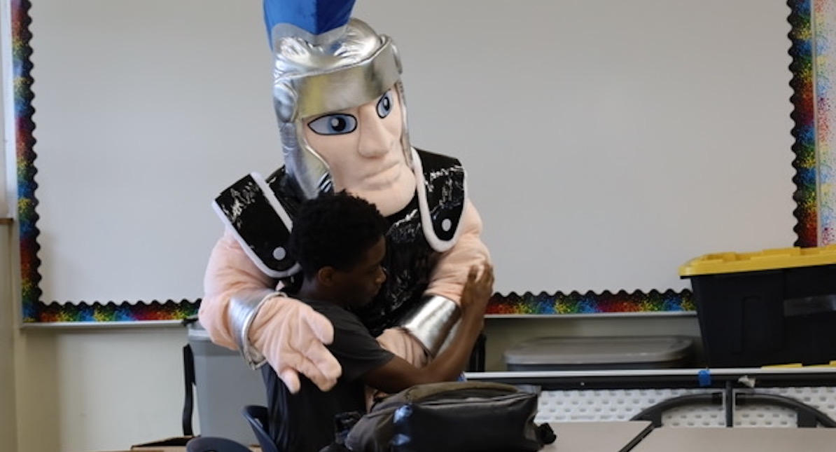 New Millennium warrior mascot hugging a high school student wearing a black shirt. Warrior mascot has silver helmet with brown vest and silver wristbands.