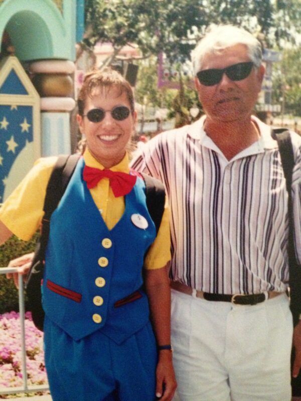 New Millennium Secondary School (NMSS) principal wears a Disneyland Toontown costume of a yellow button-down shirt with blue vest and bottoms and a bright red bow tie in front of a theme park attraction. She stands next to her dad who is wearing sunglasses, a striped polo shirt and white shorts. 
